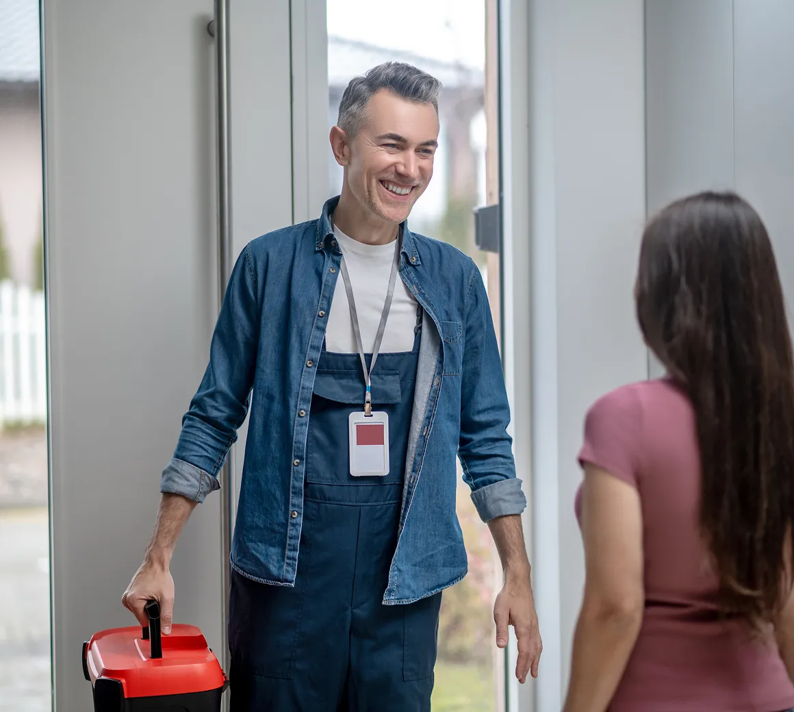 Ein lächelnder Handwerker mit blauer Arbeitskleidung und Ausweis hält einen roten Werkzeugkasten und spricht mit einer Frau in einem rosa T-Shirt im Hausflur.