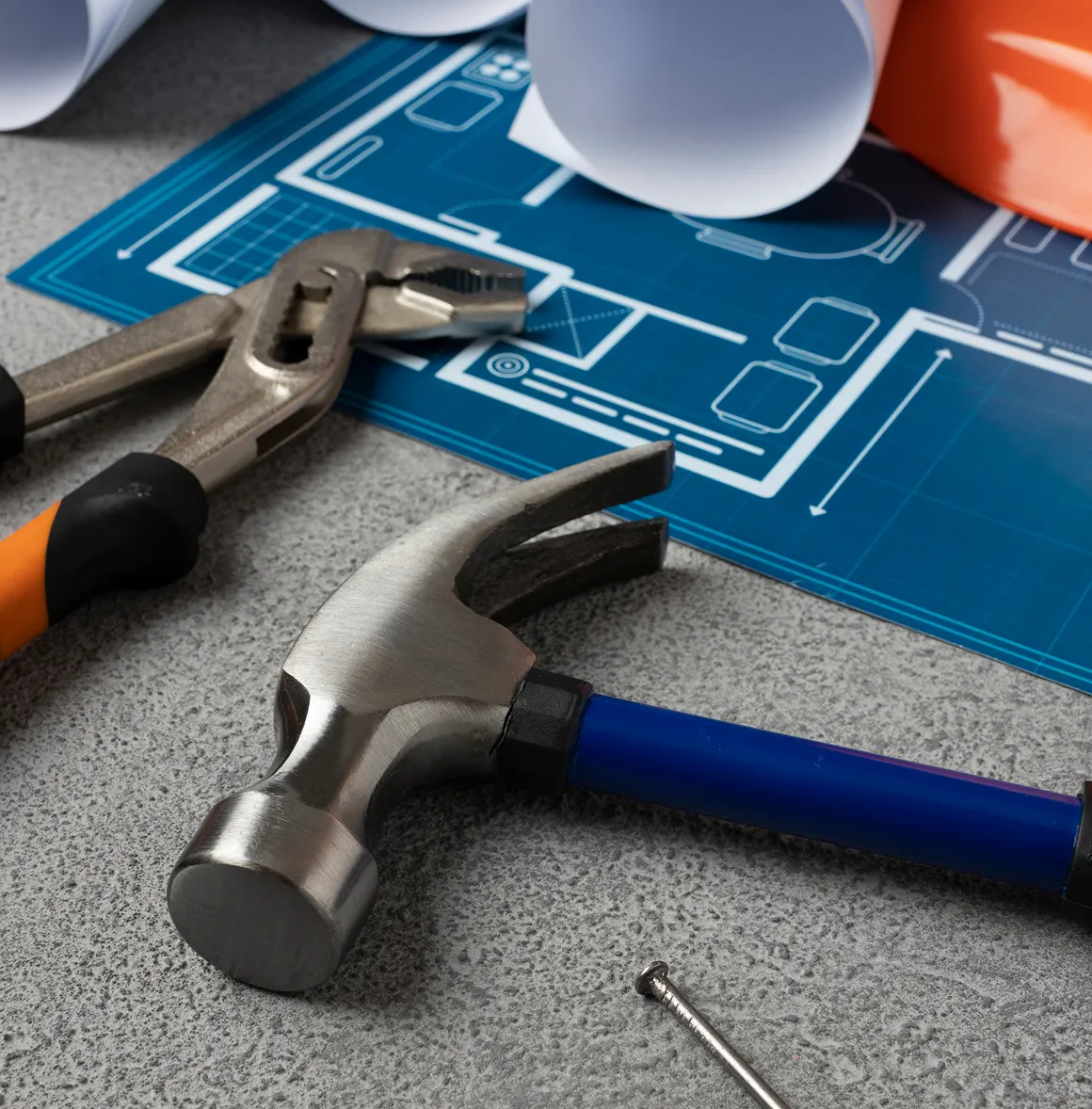 Hammer und Wasserpumpenzange liegen neben einer Bauzeichnung und einem Nagel auf einer grauen Arbeitsfläche.