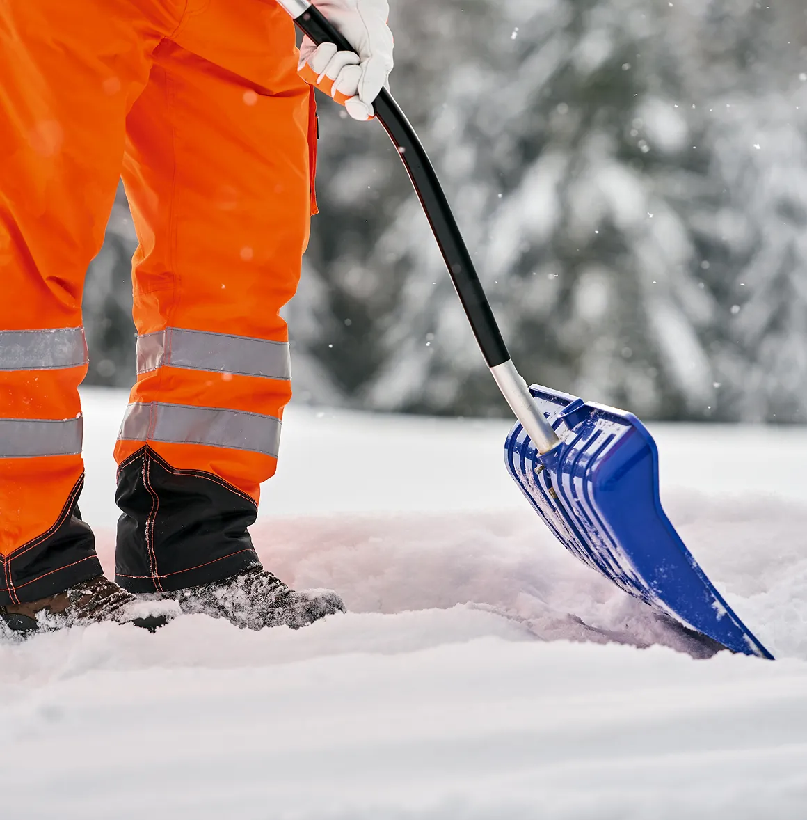 Person in orange Arbeitskleidung schaufelt Schnee mit einer blauen Schneeschaufel.