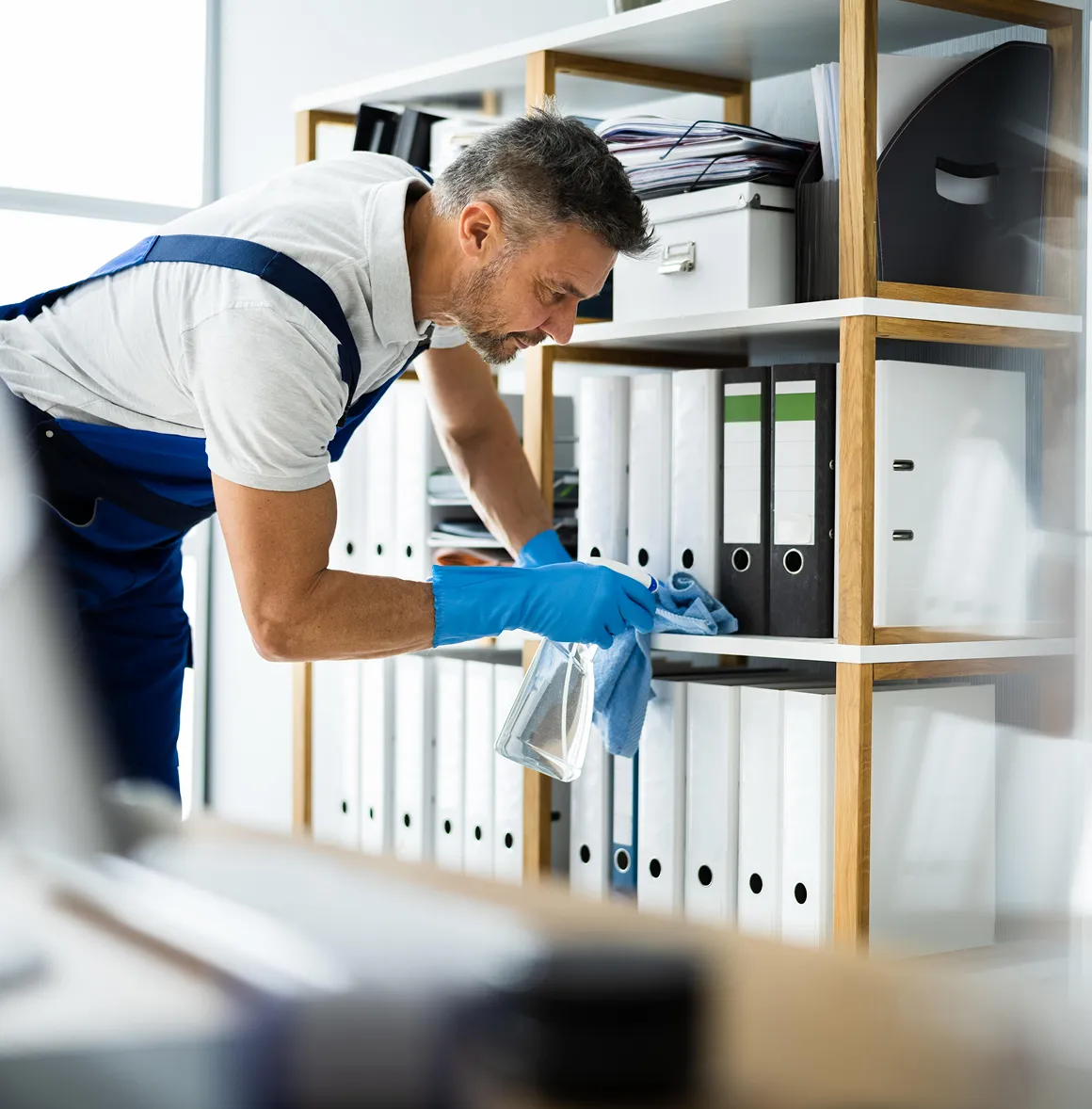 Mann mit blauen Handschuhen reinigt Regale mit Ordnern in einem Büro.
