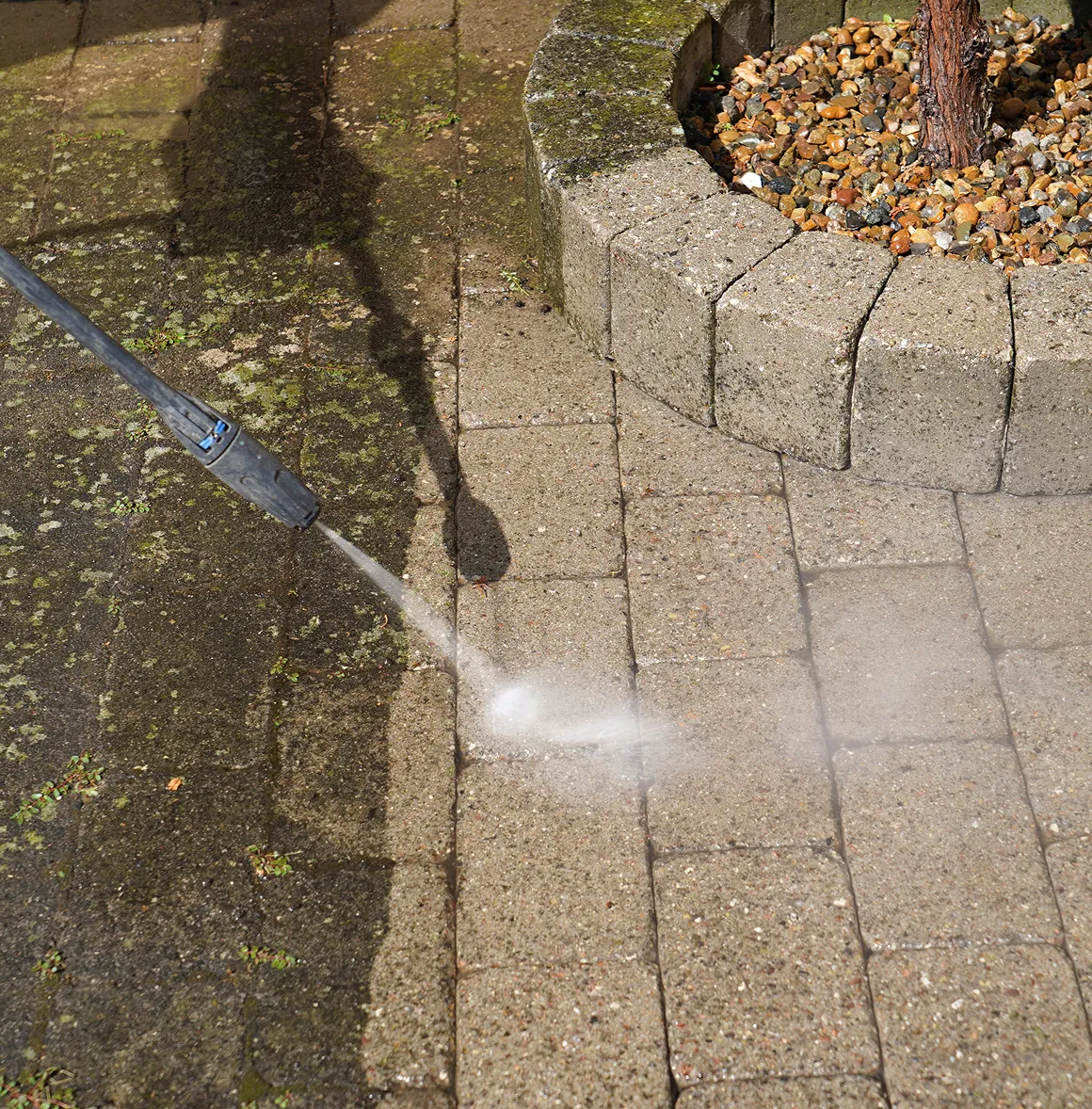 Hochdruckreiniger entfernt Schmutz und Moos von gepflastertem Boden neben einem runden Begrenzungsstein mit kleinen Steinen und einem Baumstamm.