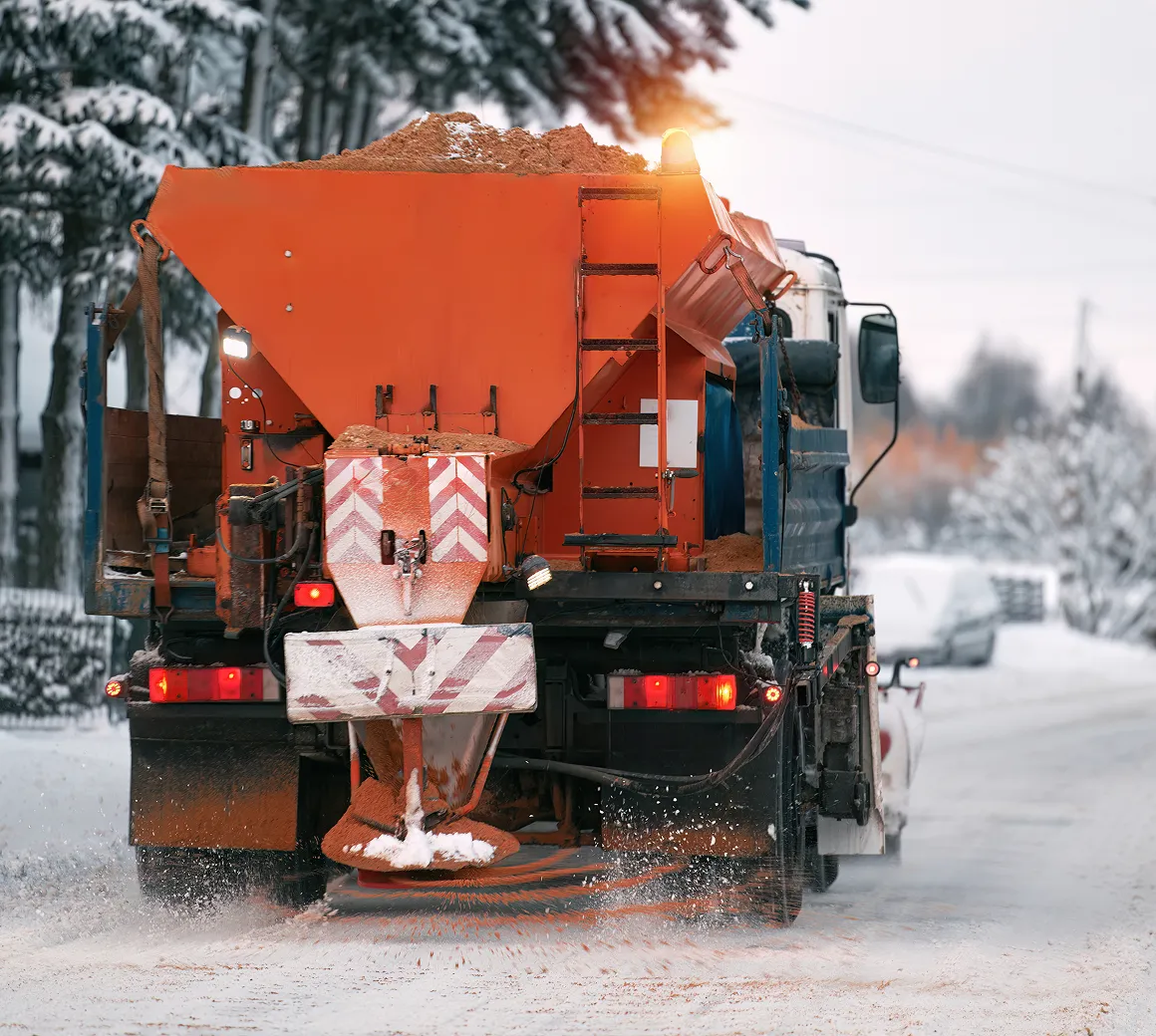 Ein Streufahrzeug verteilt Salz oder Streumittel auf einer schneebedeckten Straße in einem winterlichen Waldgebiet.