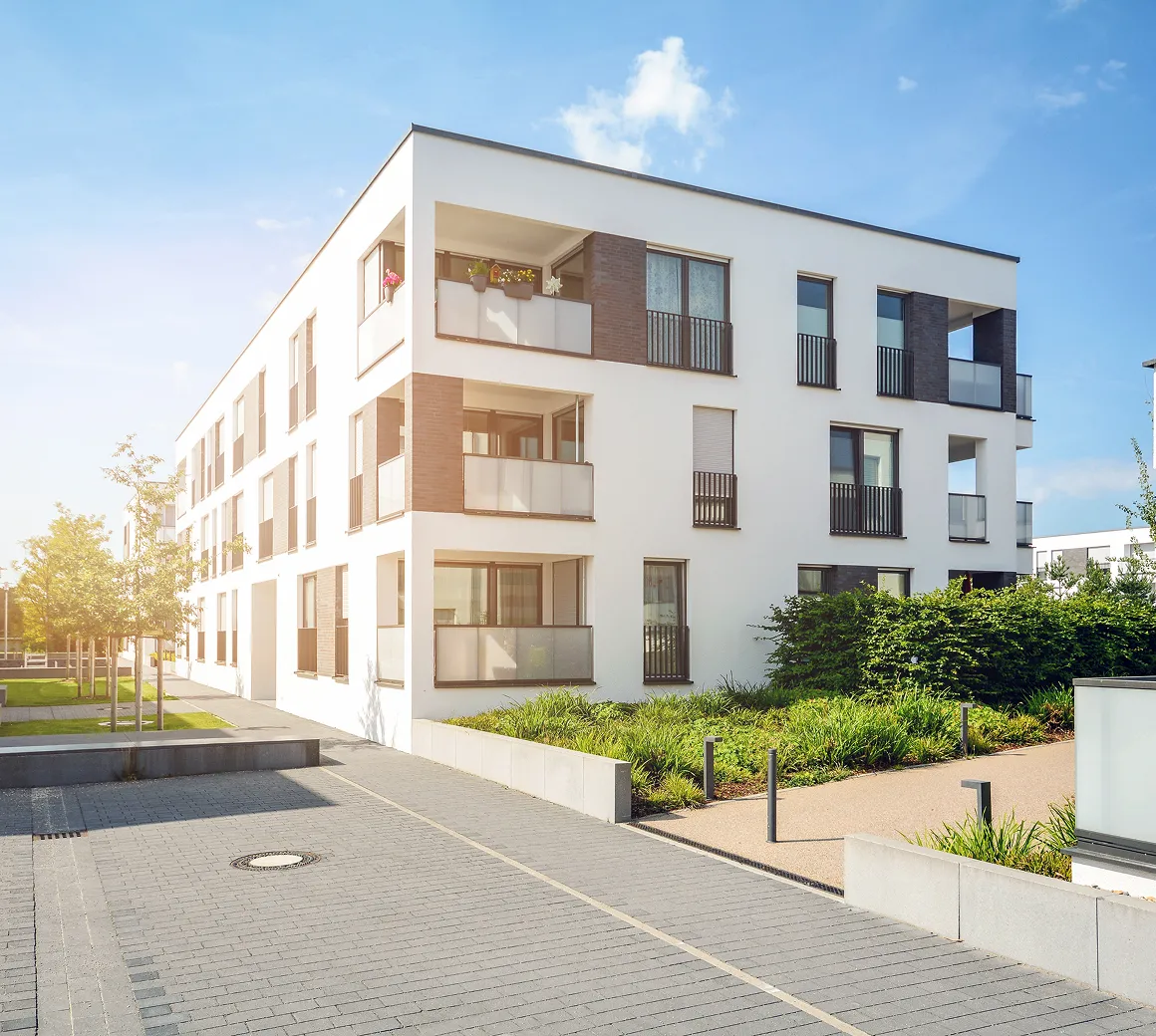 Moderne weiße Wohnhausfassade mit Balkonen, umgeben von Gehwegen, Rasenflächen und Büschen bei Sonnenschein.