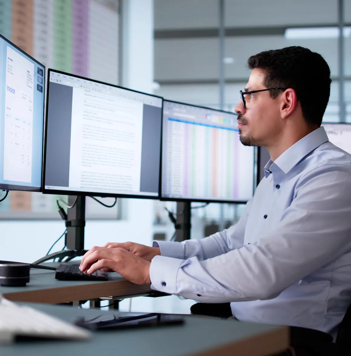 Mann mit Brille arbeitet an einem Schreibtisch mit drei Monitoren, die Dokumente und Tabellen zeigen.