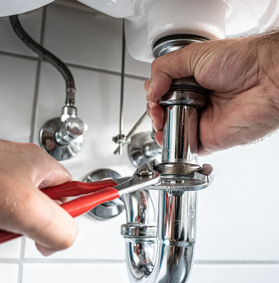 Hände ziehen mit einer Zange eine Rohrverbindung unter einem Waschbecken fest.