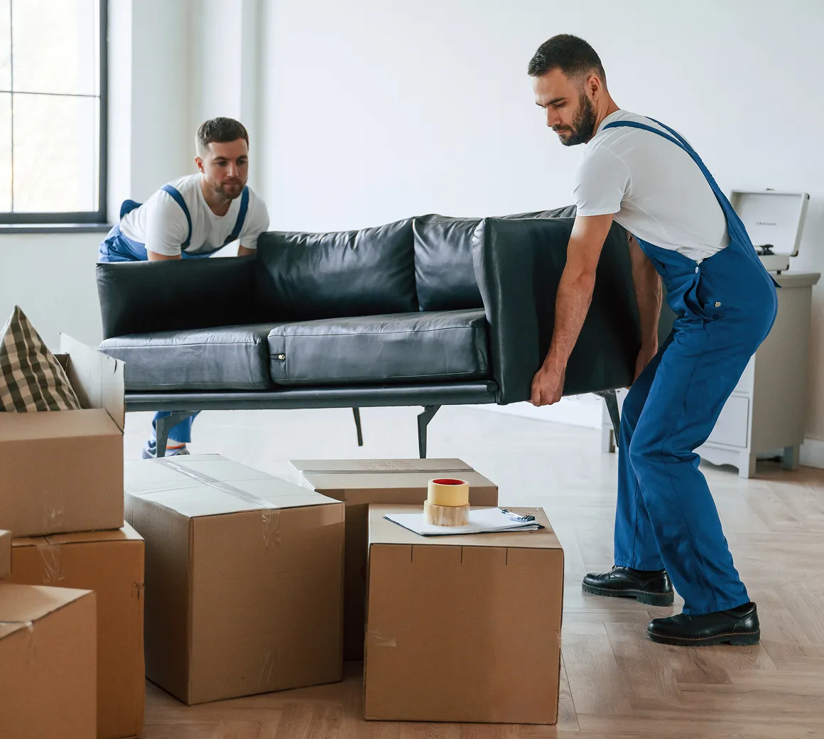 Zwei Männer in blauen Overalls heben ein schwarzes Ledersofa in einem Raum mit Kartons und einem Fenster.