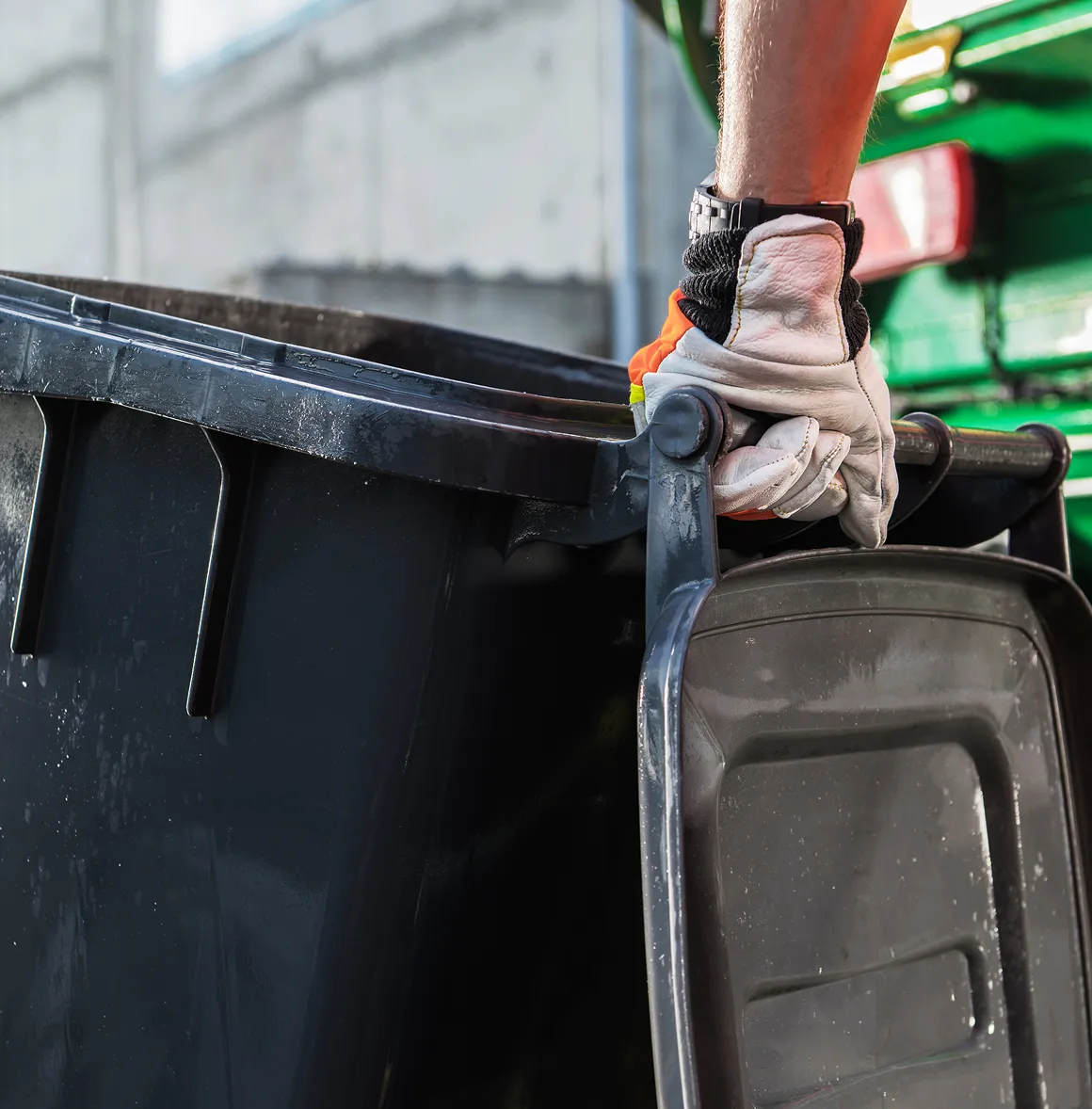 Hand mit Arbeitshandschuh hebt den Deckel eines schwarzen Müllcontainers an.