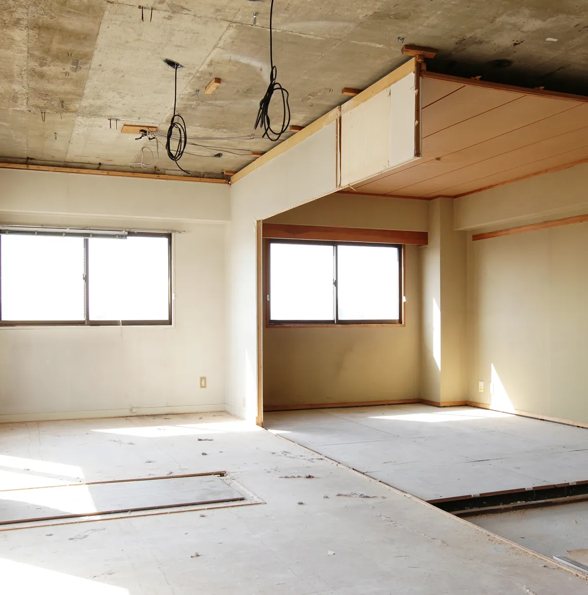 Leerer Raum mit zwei Fenstern, unvollständigem Boden und sichtbarer Betondecke mit herunterhängenden Kabeln.