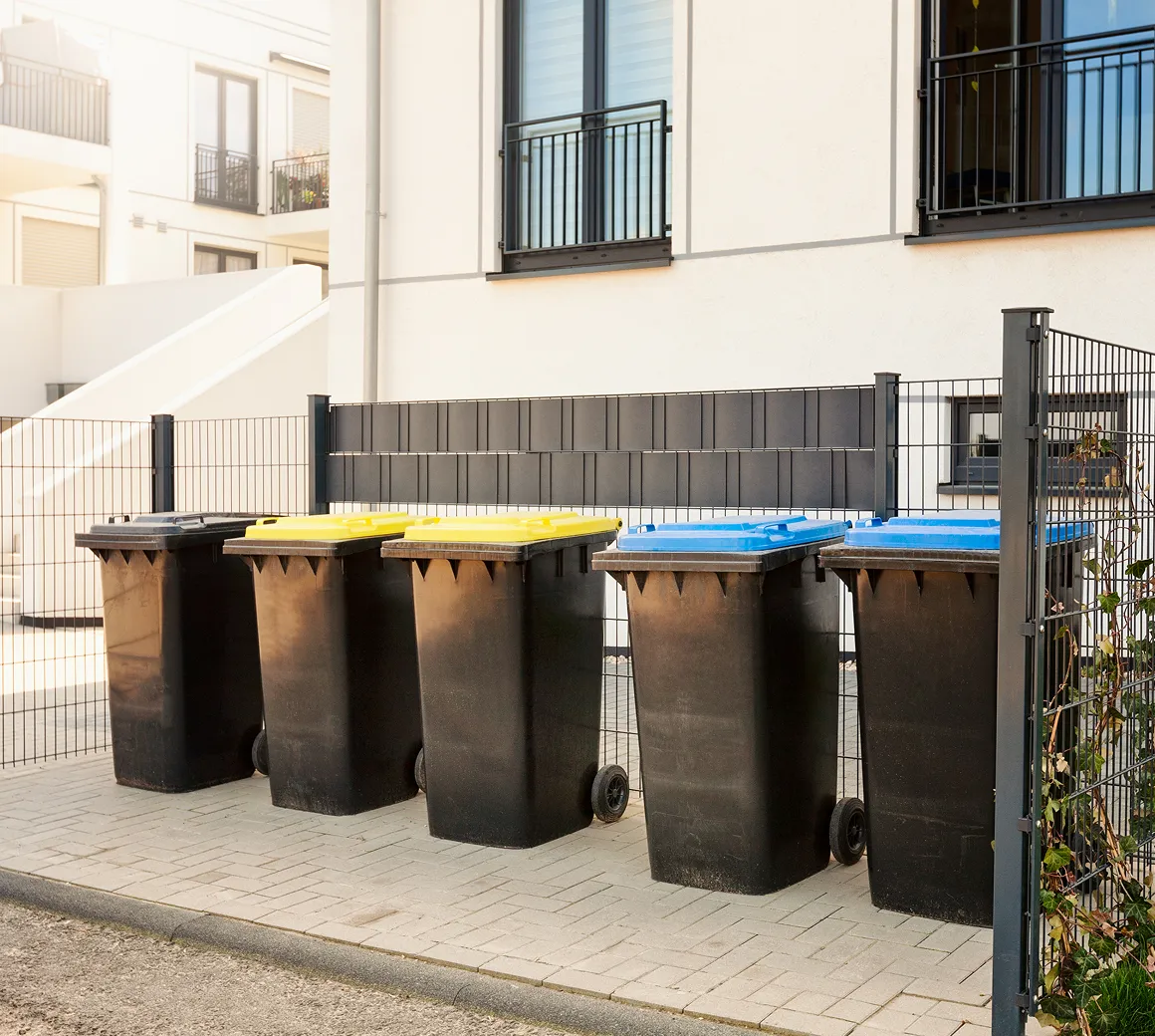 Fünf Mülltonnen mit schwarzen Behältern und Deckeln in Gelb, Blau und Schwarz stehen nebeneinander auf einem gepflasterten Bereich vor einem Zaun und einem weißen Gebäude.