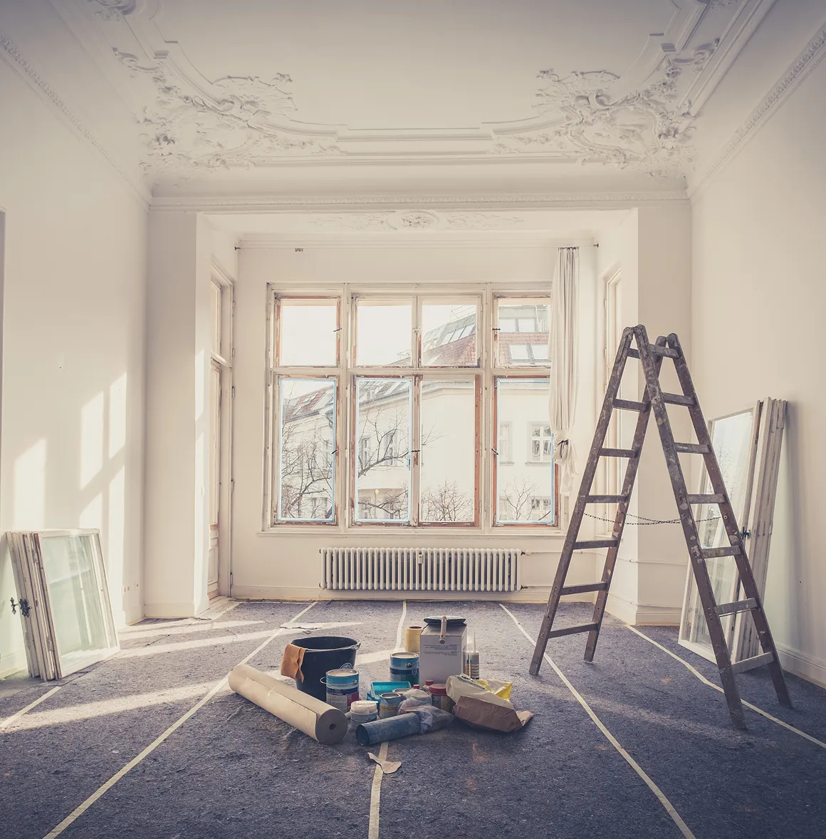 Helles Zimmer mit hohen Fenstern und dekorativer Decke, in Renovierung mit Leiter, Farbeimern und Bauutensilien.