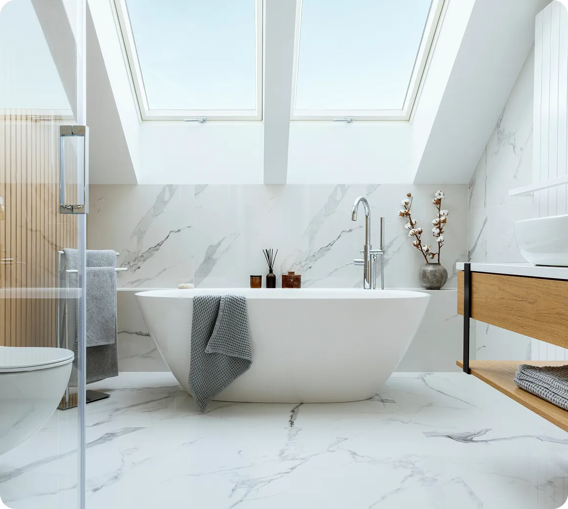 Modernes Badezimmer mit freistehender Badewanne unter zwei großen Dachfenstern, hellgrauem Handtuch und grauem Bademantel auf weißem Marmorboden.