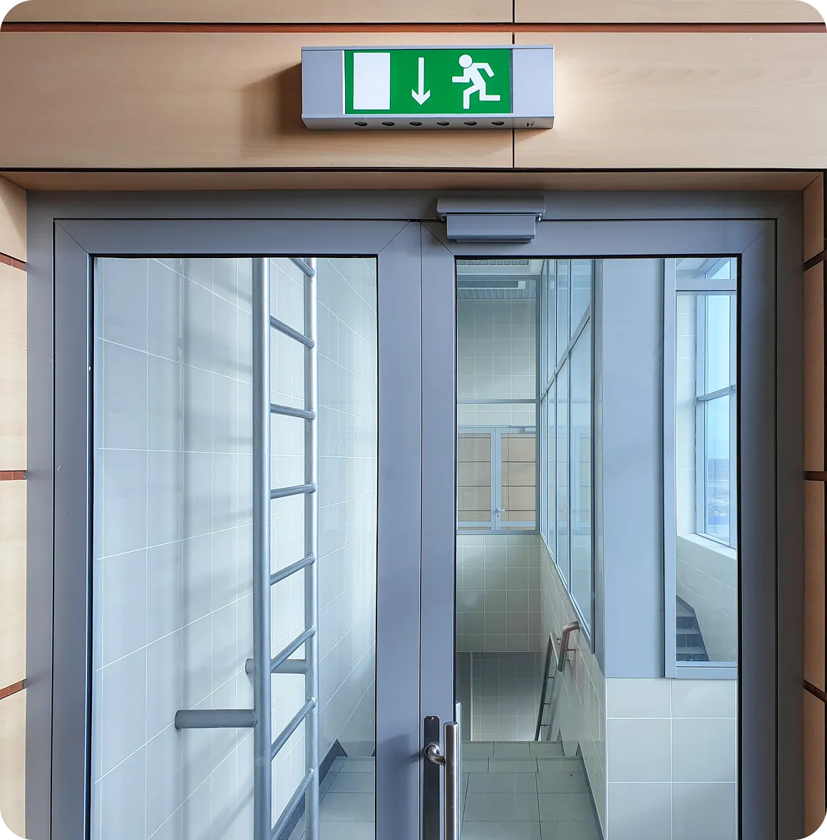 Doppeltür mit Glaseinsätzen vor einem Treppenhaus und einem grünen Notausgangsschild mit einer nach unten zeigenden Pfeil- und Fluchtsymbolik.