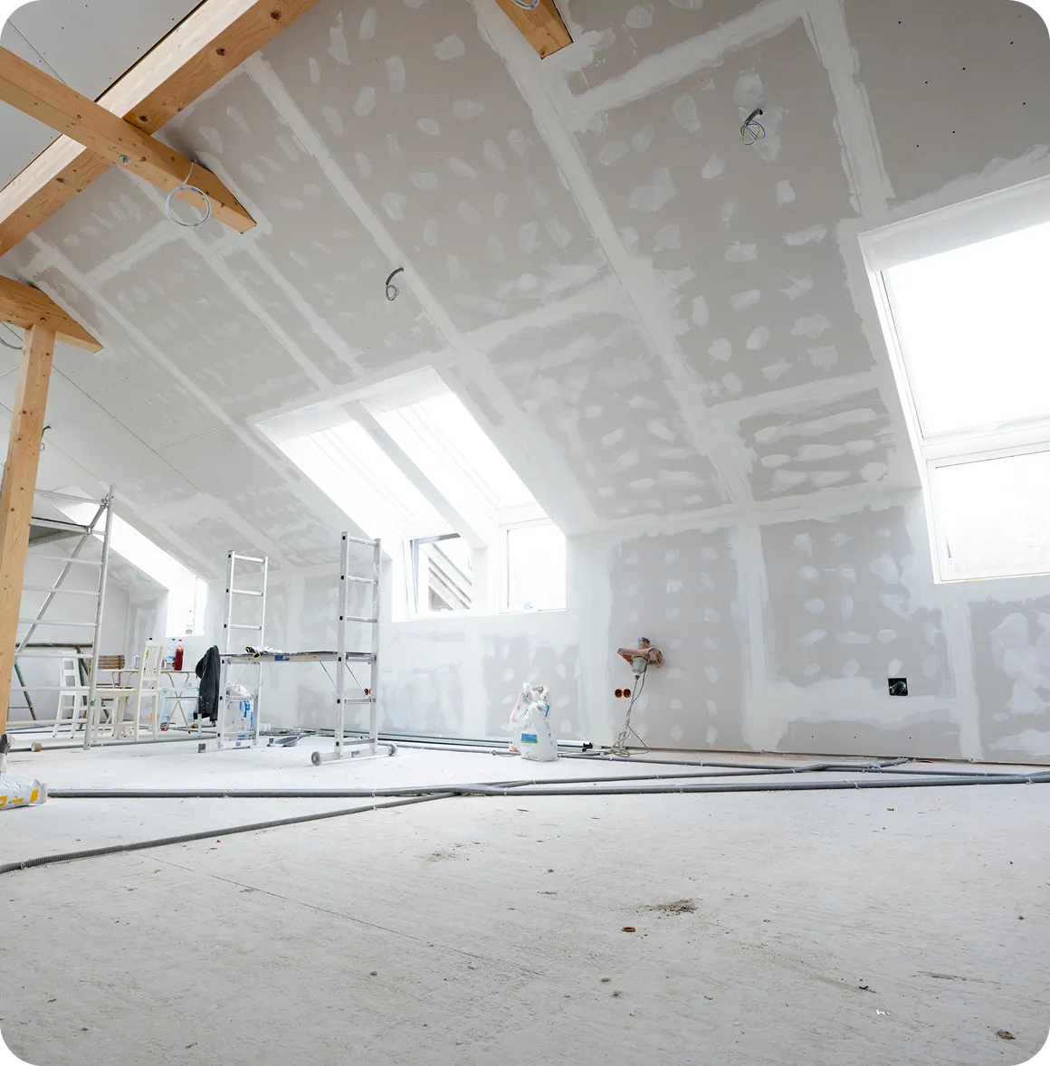 Großer, heller Raum im Rohbau mit Gipskartonwänden, Holzträgern, Leitungen auf dem Boden und zwei großen Dachfenstern.