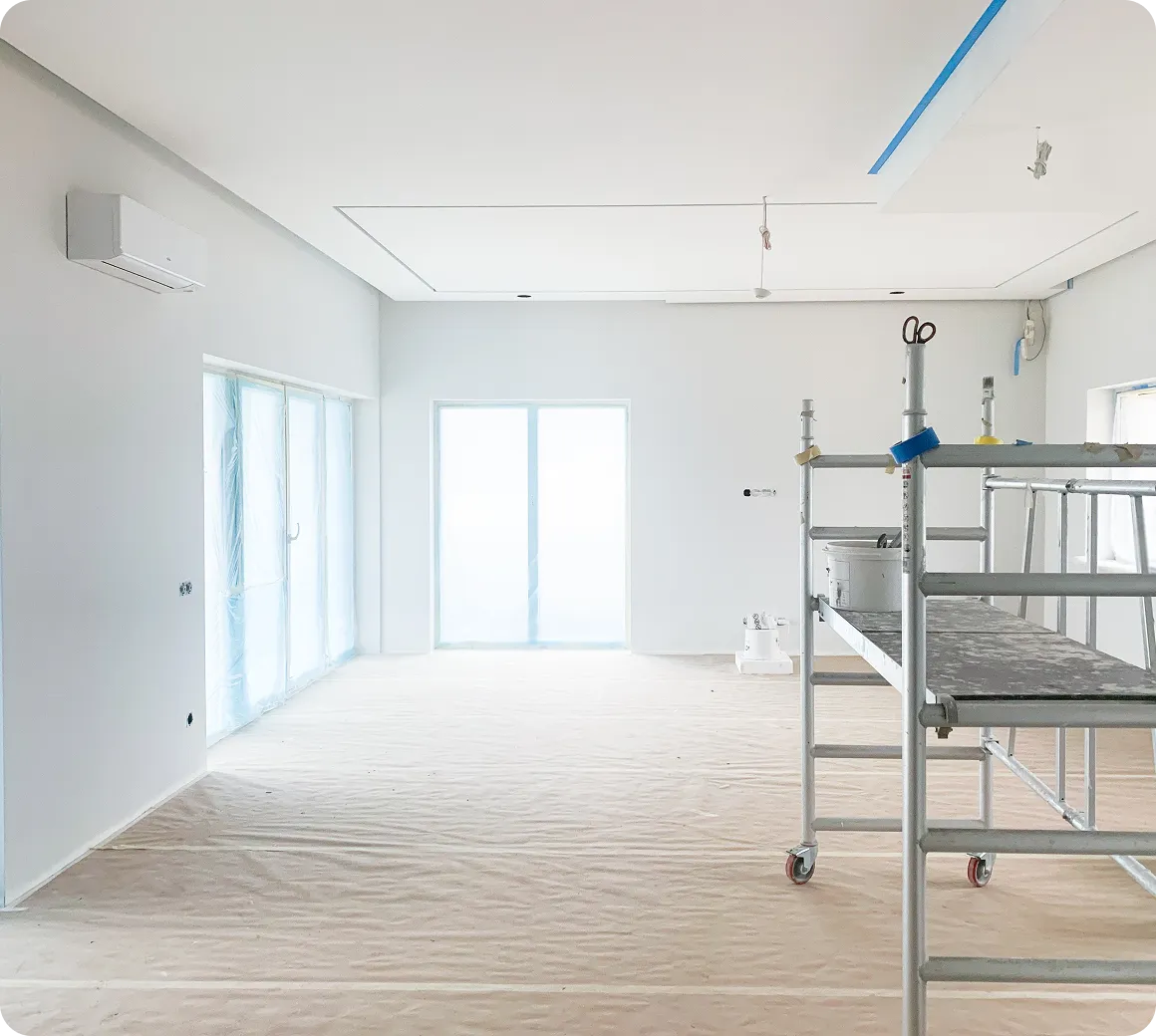 Helles, unmöbliertes Zimmer mit eingehüllten Fenstern und einer Rollgerüstplattform für Renovierungsarbeiten.