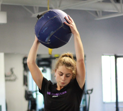 Young woman in athletic wear lifts a large blue medicine ball overhead in a gym. She appears focused and determined, surrounded by fitness equipment.