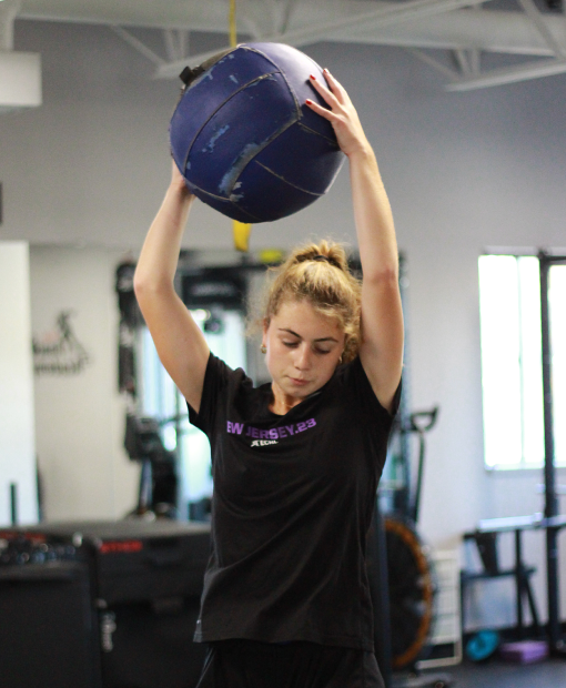Young woman in athletic wear lifts a large blue medicine ball overhead in a gym. She appears focused and determined, surrounded by fitness equipment.