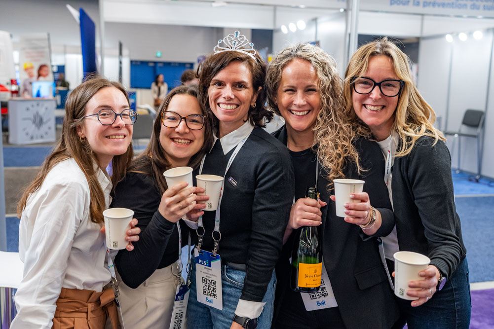 Five smiling women holding cups and a bottle, one wearing a tiara, at an indoor event.