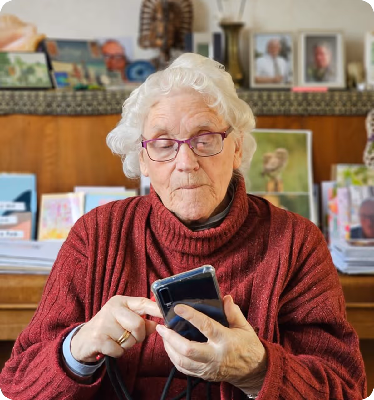 Elderly woman wearing glasses and a red sweater using a smartphone indoors.