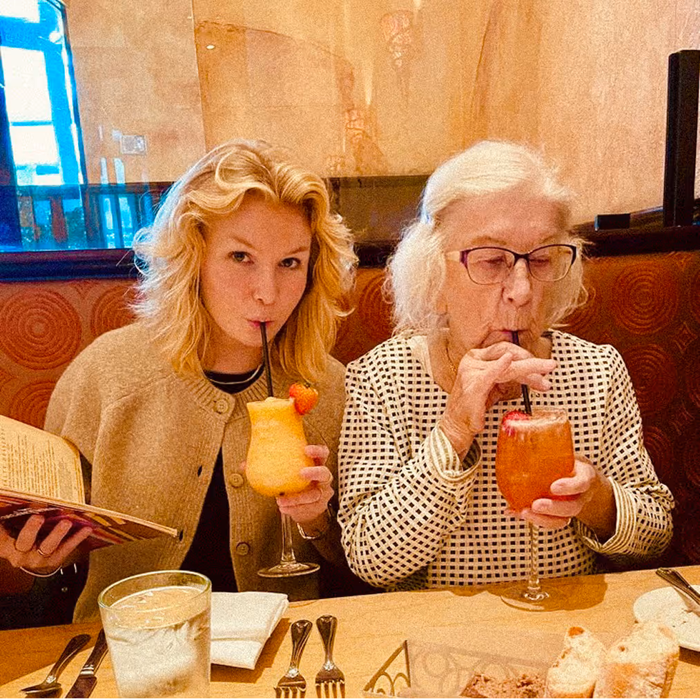 Older woman in a purple fleece jacket and a younger woman wearing glasses, both seated and wearing seatbelts inside a car, with the younger woman smiling and taking a selfie.