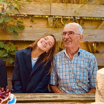 Smiling elderly man with glasses and plaid shirt sitting next to a young woman with long hair wearing a black blazer, both seated at a wooden table outdoors.