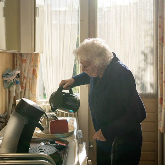 A young woman in a black blazer smiling and leaning her head toward an elderly man in glasses and a checked shirt sitting beside her outdoors.