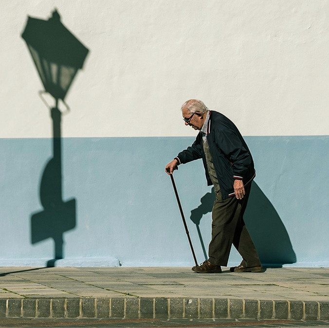 A young woman in a black blazer smiling and leaning her head toward an elderly man in glasses and a checked shirt sitting beside her outdoors.