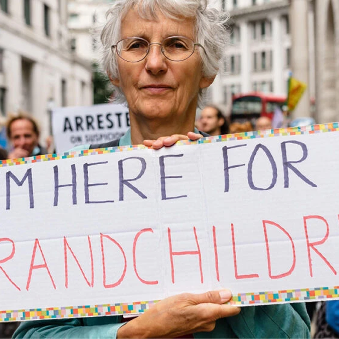 A young woman in a black blazer smiling and leaning her head toward an elderly man in glasses and a checked shirt sitting beside her outdoors.