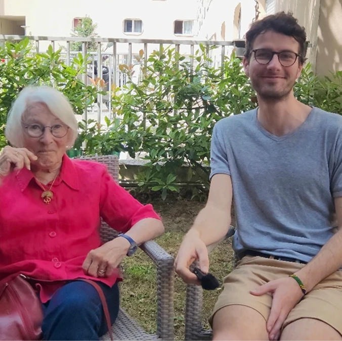 A young woman in a black blazer smiling and leaning her head toward an elderly man in glasses and a checked shirt sitting beside her outdoors.