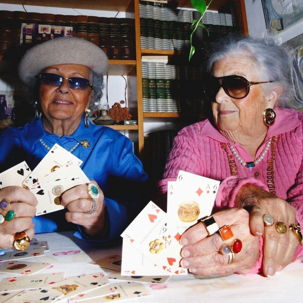 A young woman in a black blazer smiling and leaning her head toward an elderly man in glasses and a checked shirt sitting beside her outdoors.