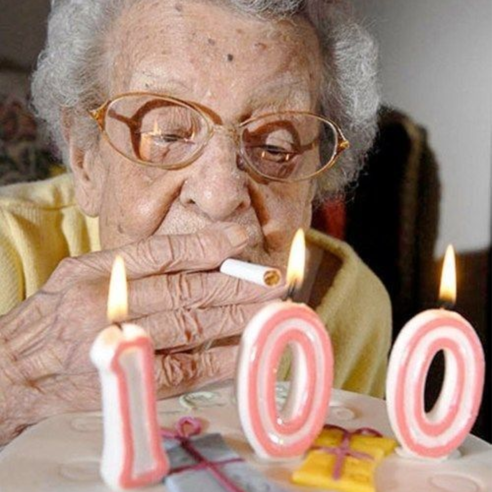 A young woman in a black blazer smiling and leaning her head toward an elderly man in glasses and a checked shirt sitting beside her outdoors.