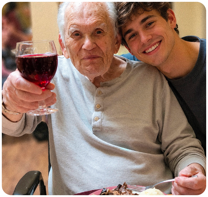 Smiling elderly man with glasses and plaid shirt sitting next to a young woman with long hair wearing a black blazer, both seated at a wooden table outdoors.
