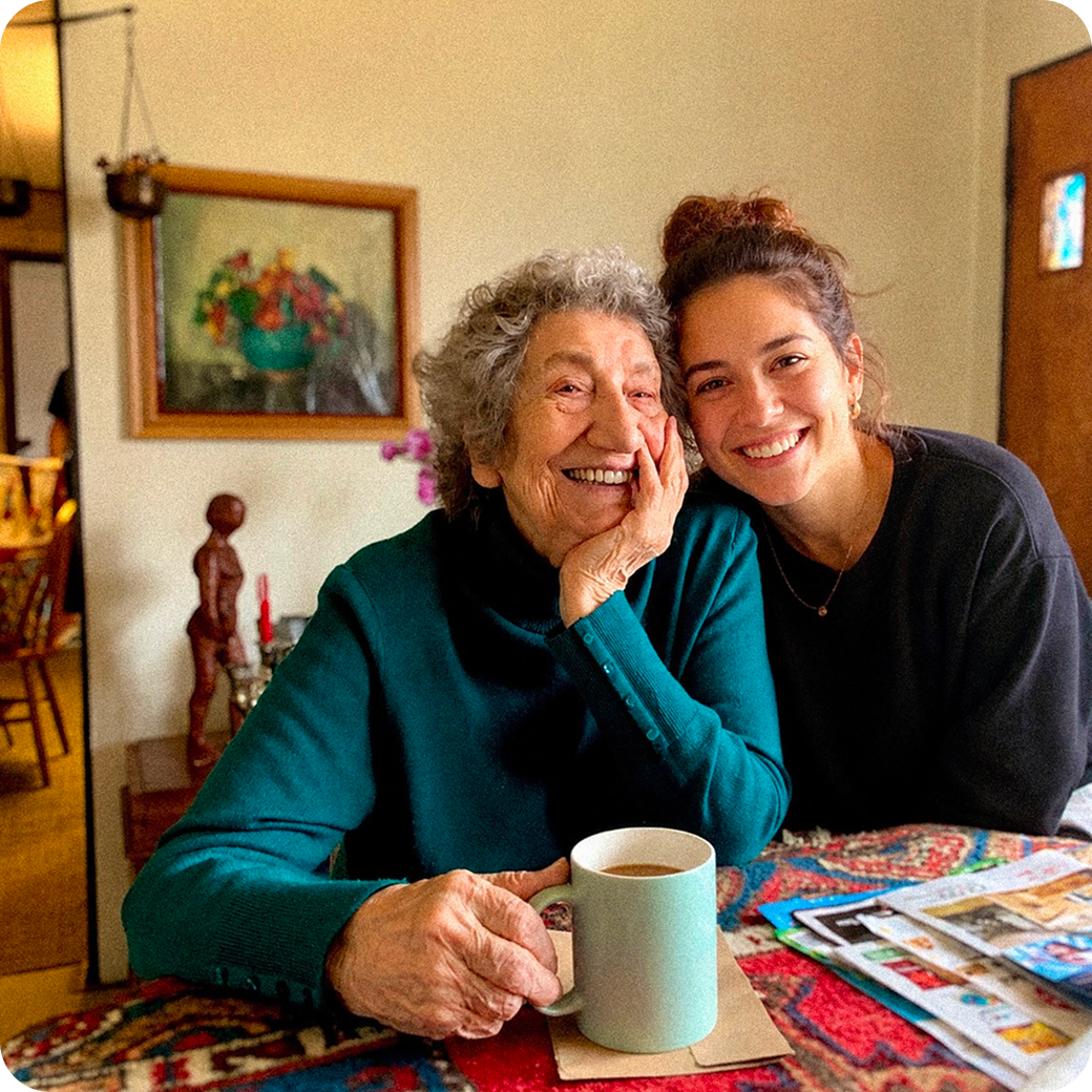 Smiling elderly man with glasses and plaid shirt sitting next to a young woman with long hair wearing a black blazer, both seated at a wooden table outdoors.