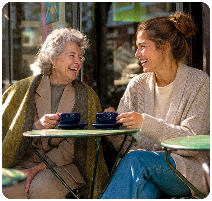 Smiling elderly man with glasses and plaid shirt sitting next to a young woman with long hair wearing a black blazer, both seated at a wooden table outdoors.