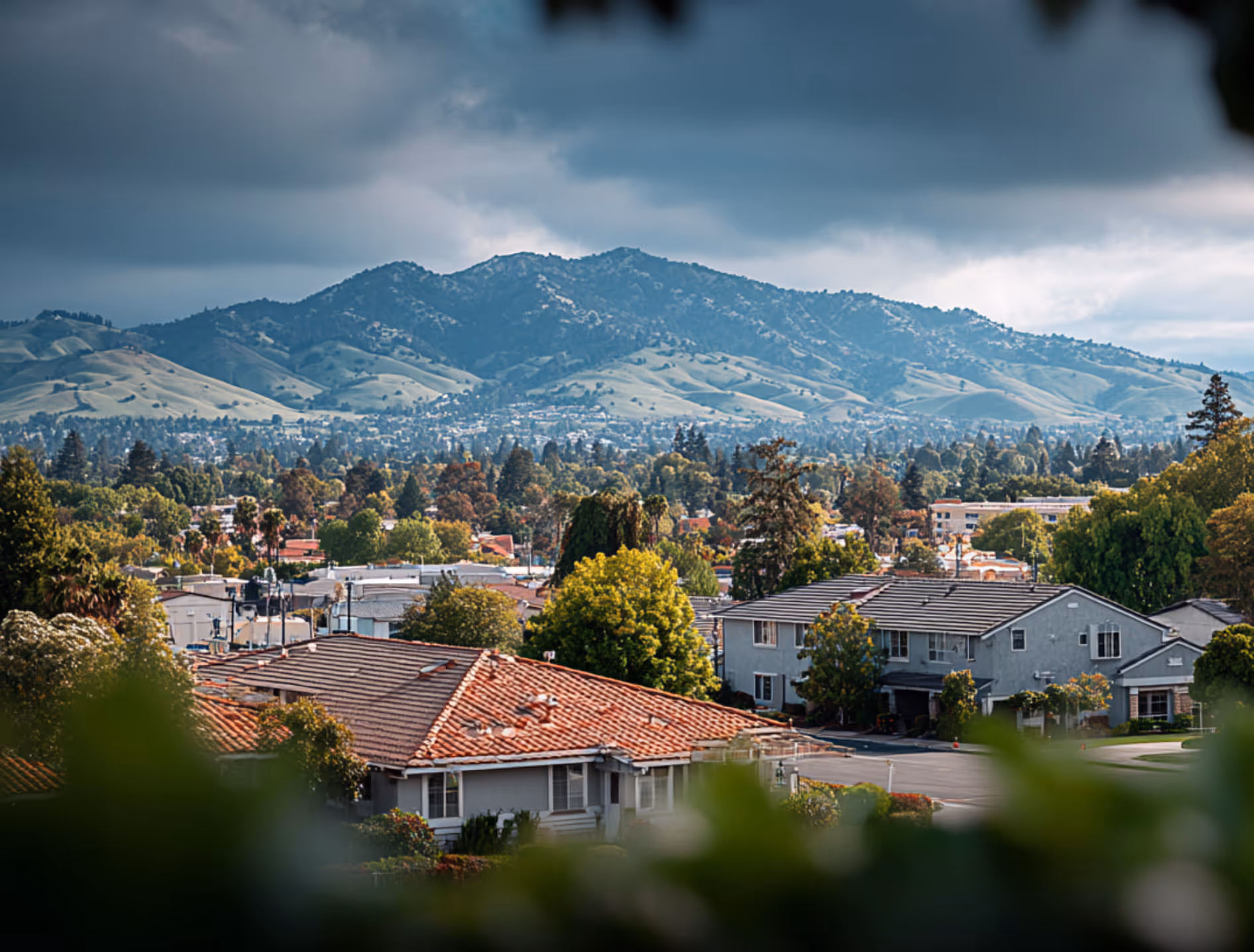 Suburban neighborhood with houses and trees in the foreground, set against a backdrop of green hills under a cloudy sky.