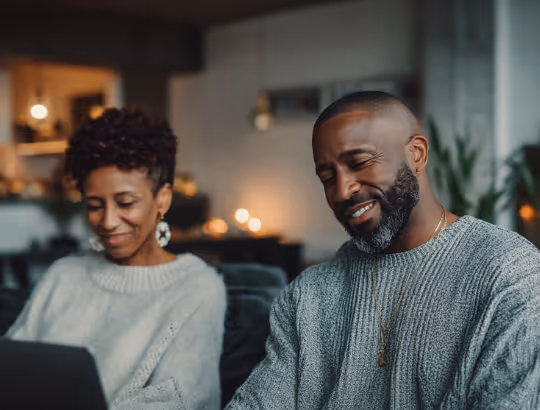 Smiling man and woman sitting together indoors, both wearing knitted sweaters and looking at a laptop.