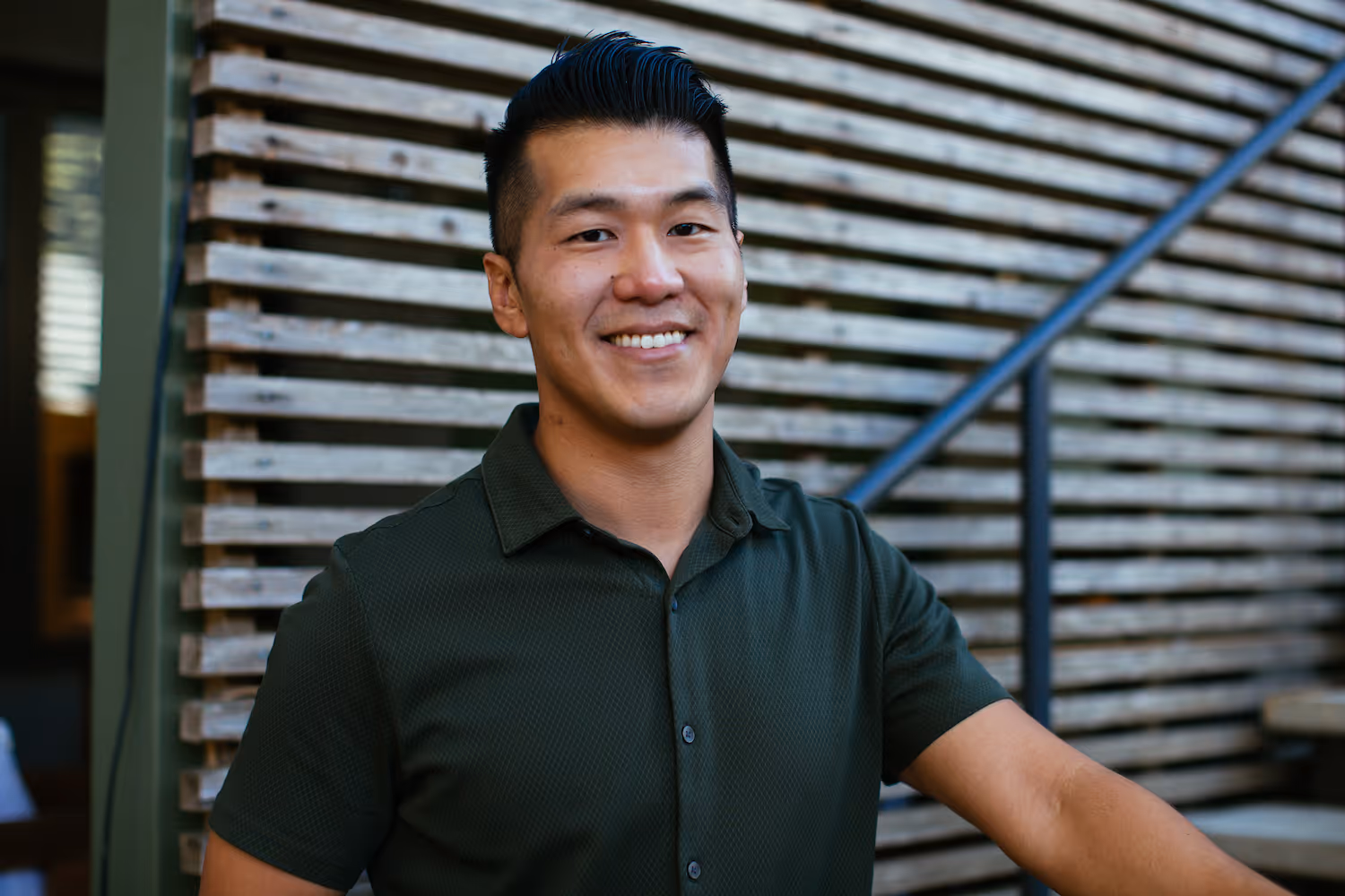 Smiling man with short black hair wearing a dark green shirt standing in front of a wooden slatted wall with handrail.