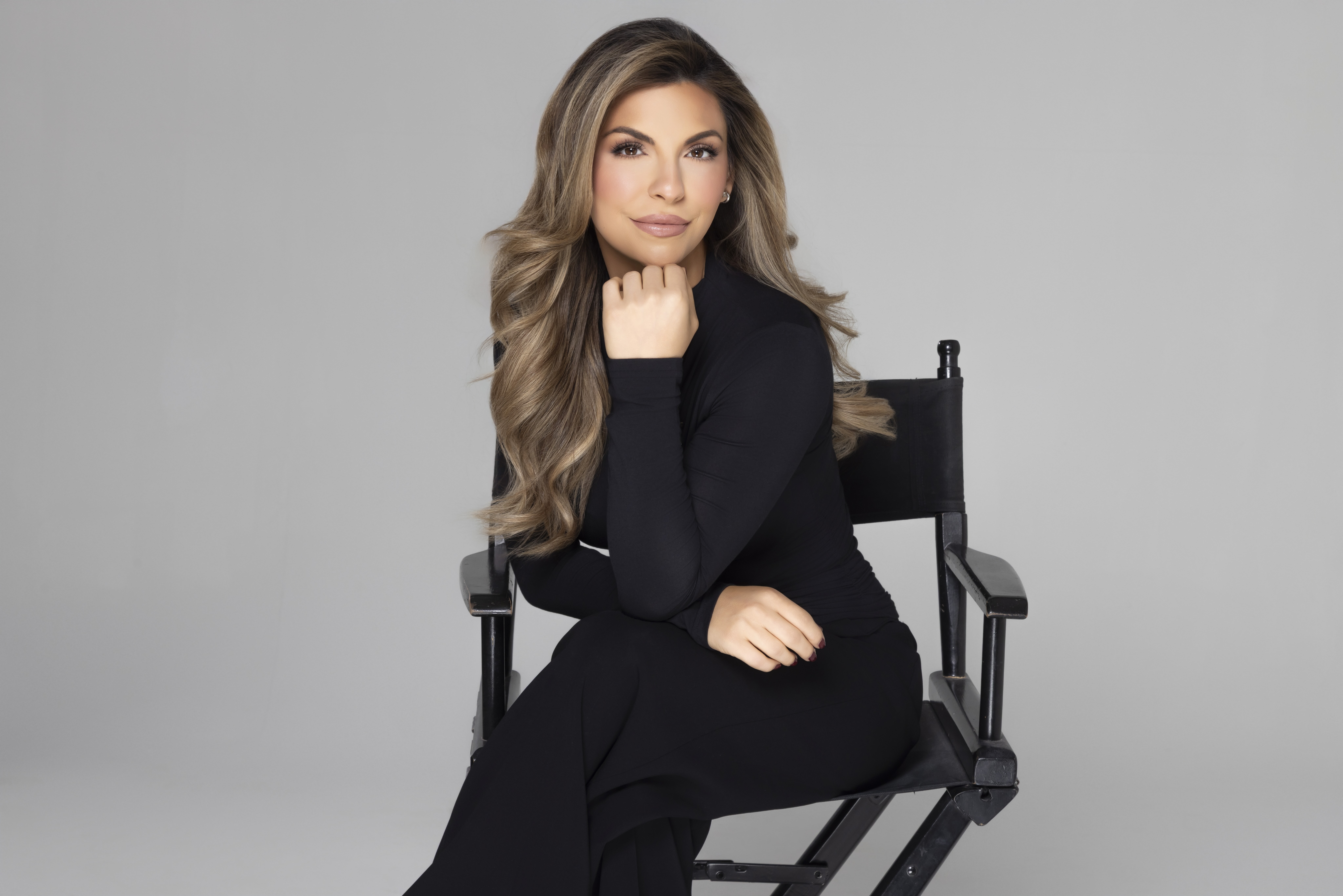 Woman with long wavy hair wearing a black outfit sitting on a director's chair against a plain gray background.