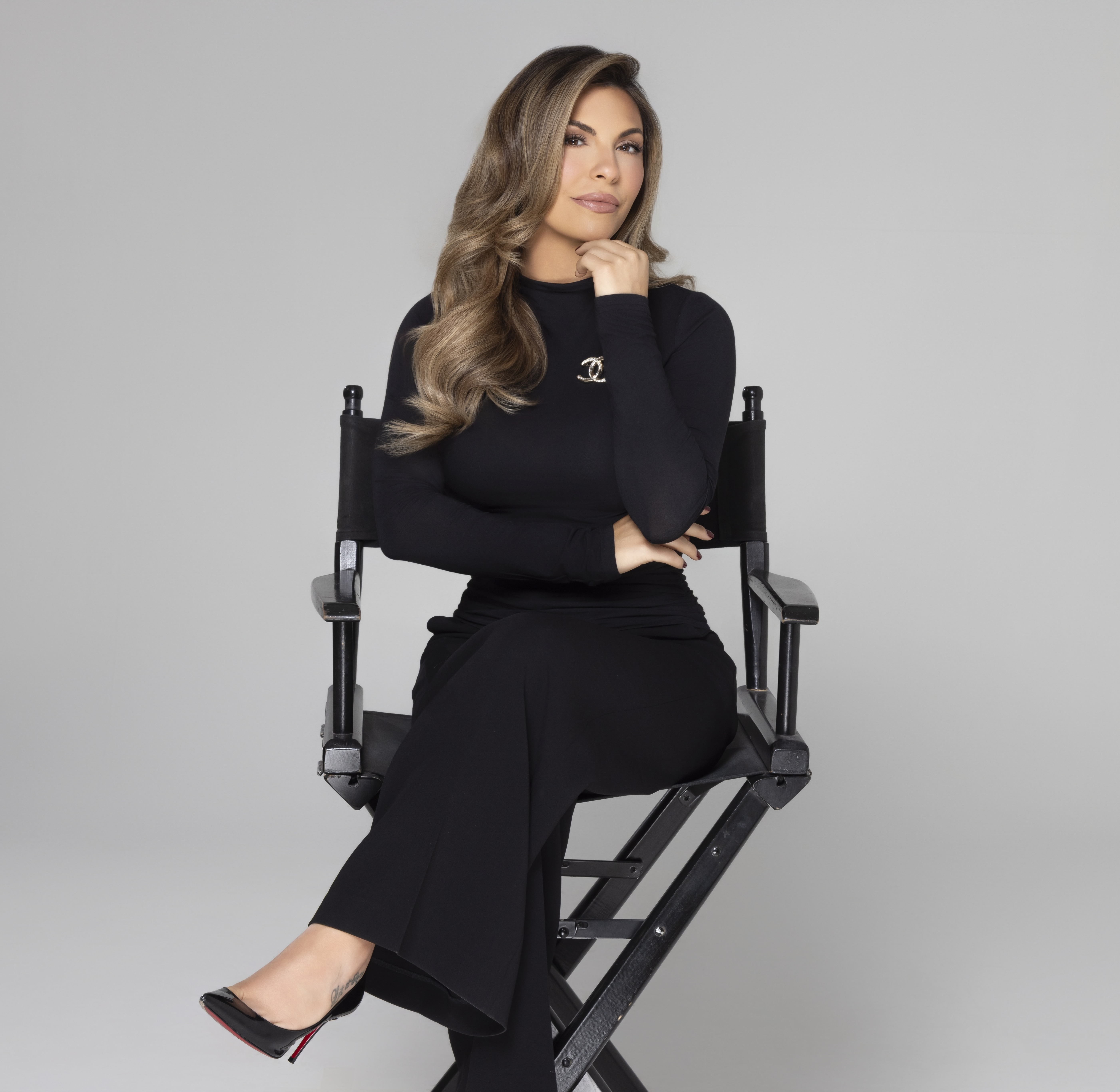 Woman with long wavy hair wearing a black dress and high heels sitting cross-legged on a director's chair against a gray background.