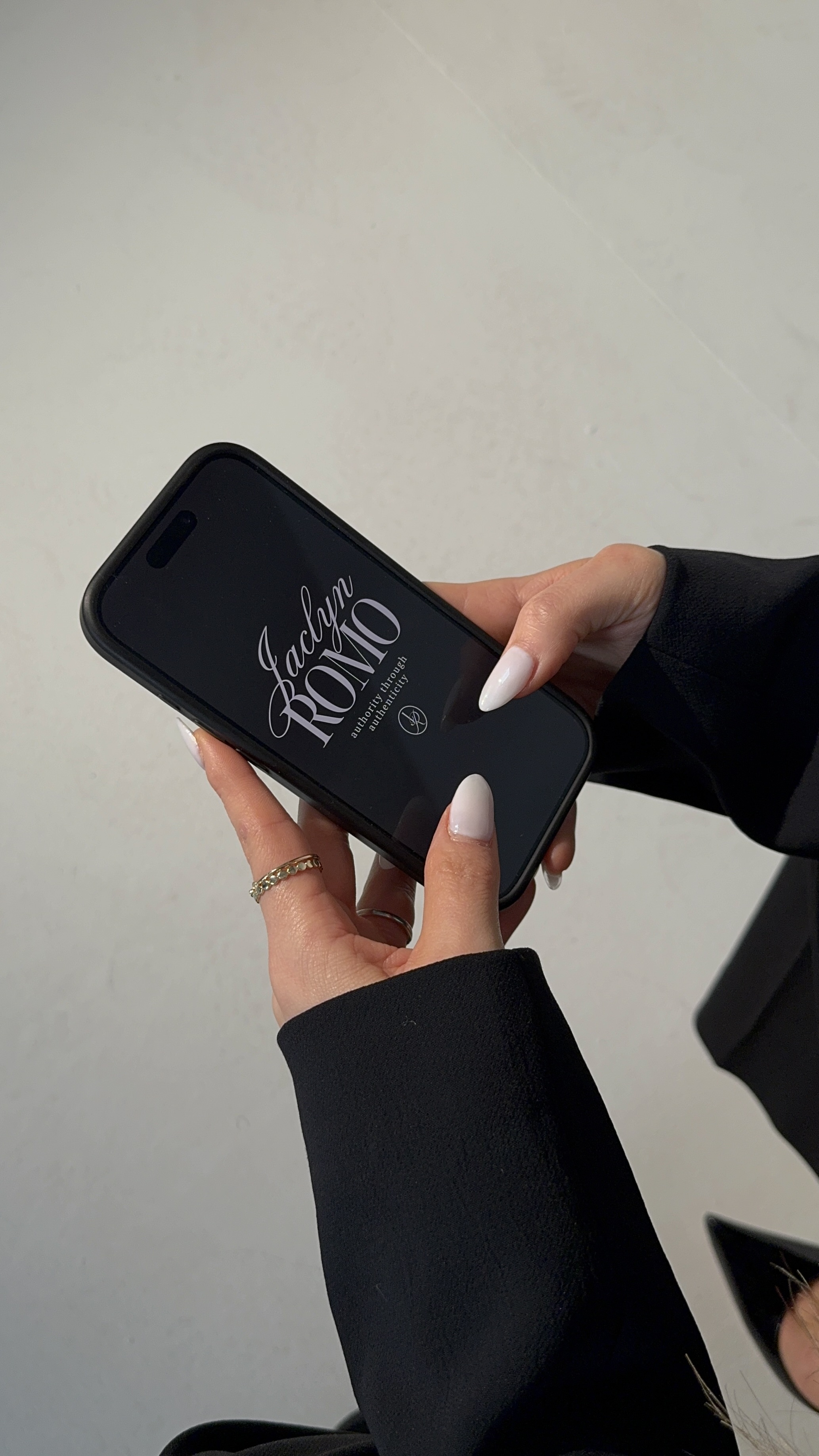 Hands with white nail polish holding a black smartphone displaying 'Jaclym ROMO authority through authenticity' on the screen.