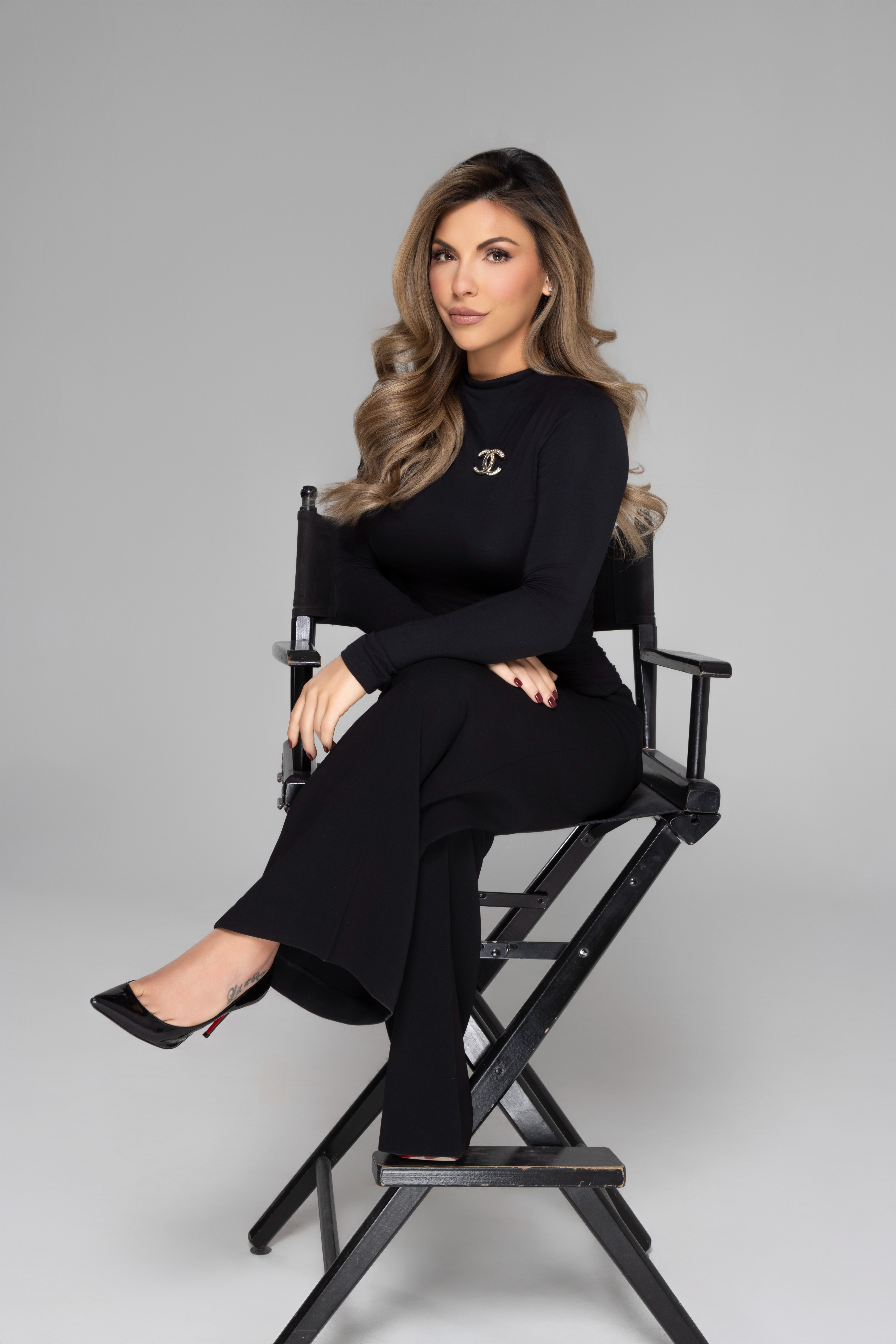 Woman with long wavy hair wearing all black, sitting cross-legged on a black director's chair against a gray background.