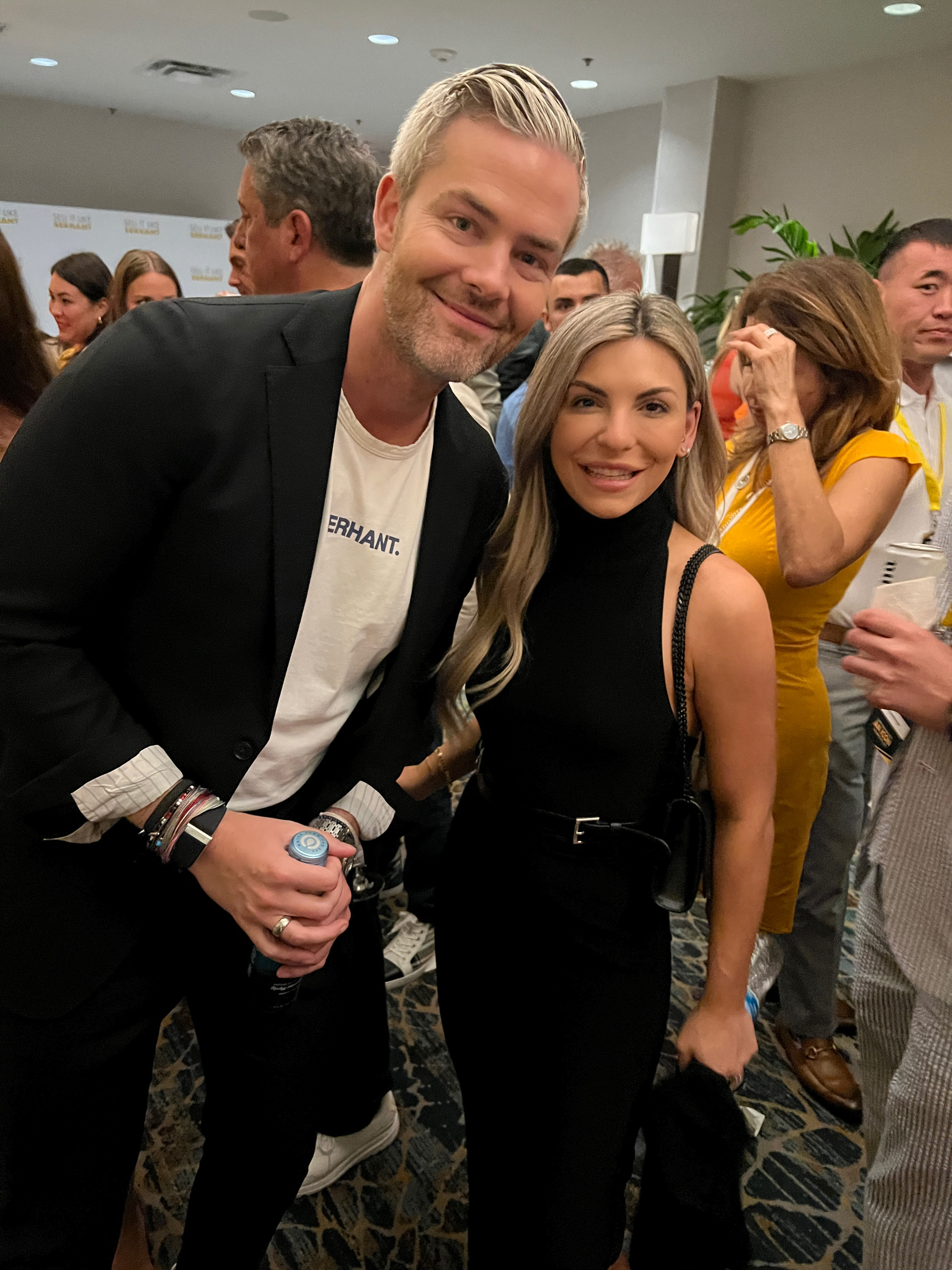 A smiling man with blond hair and a black blazer posing next to a woman with long blond hair wearing a sleeveless black dress at an indoor social event.
