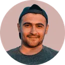 Young man with short dark hair wearing a backwards black cap and a dark shirt, smiling slightly against a muted pink background.