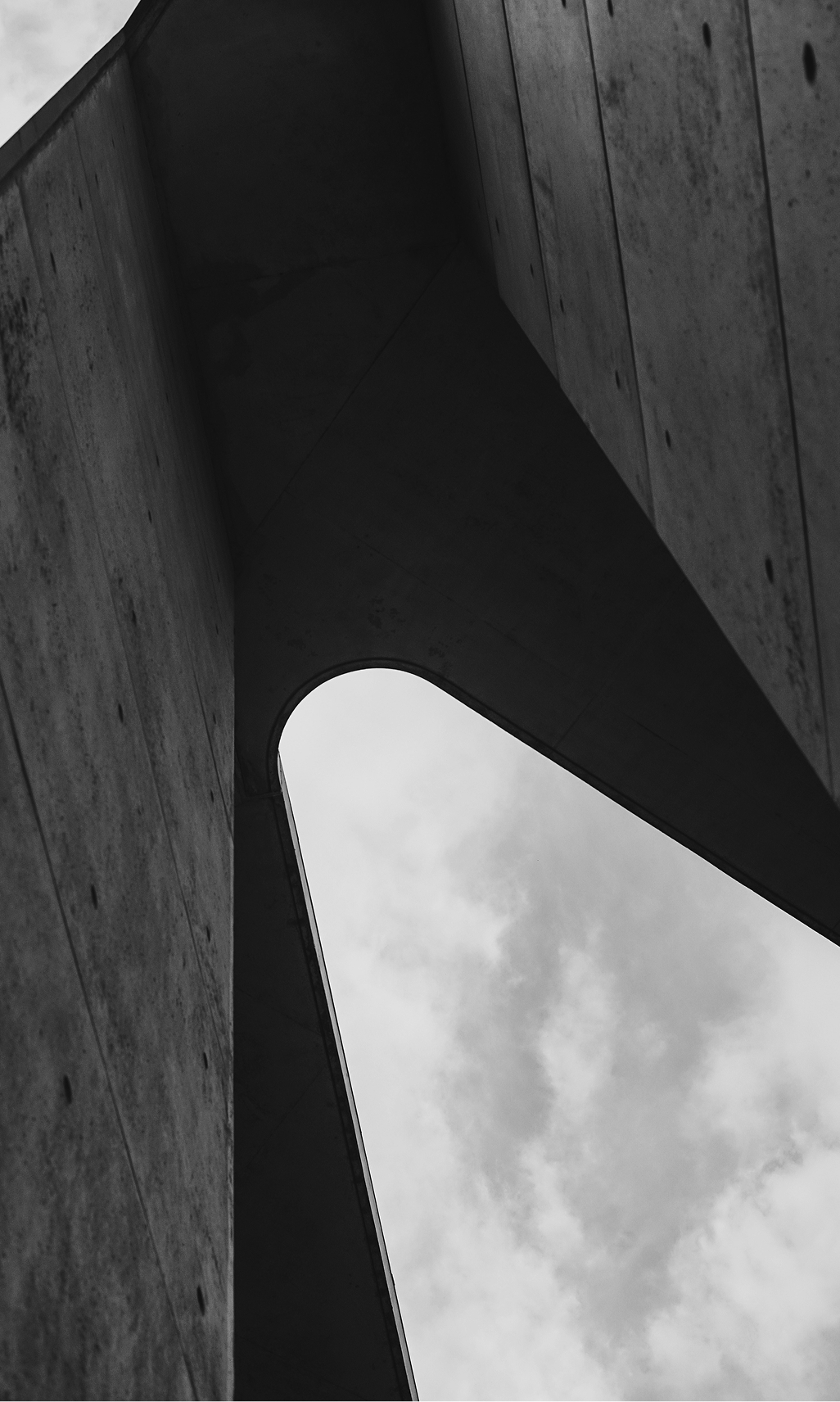 Black and white photo looking up at a modern concrete building with an open curved gap showing cloudy sky.