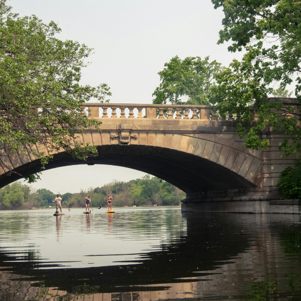 Three people paddleboarding under a stone bridge on a calm river surrounded by green trees.