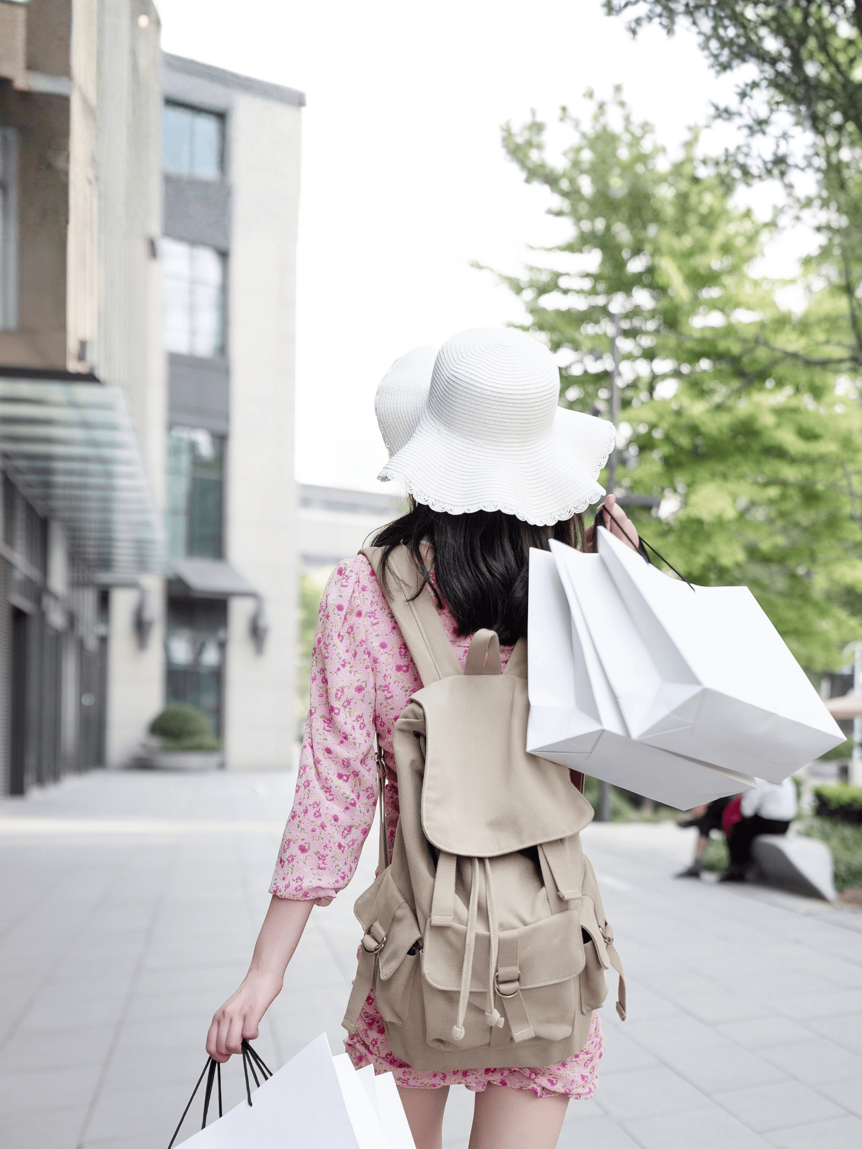 Woman wearing a white sun hat and pink floral dress carrying white shopping bags and a beige backpack in an urban area.