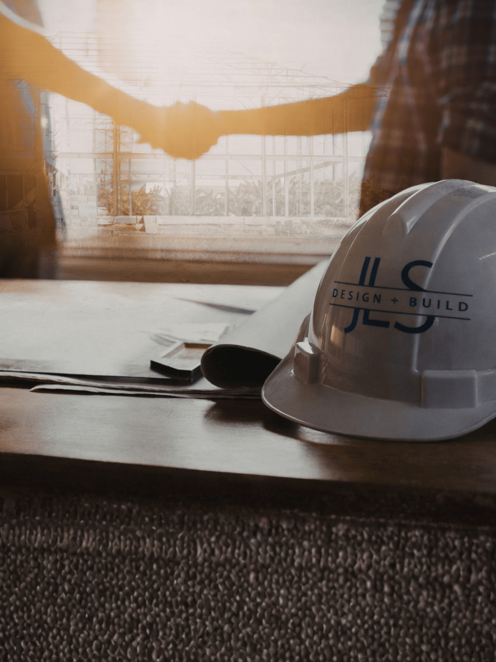 White construction helmet with JLS Design + Build logo on a table near rolled blueprints with two people shaking hands in the background.