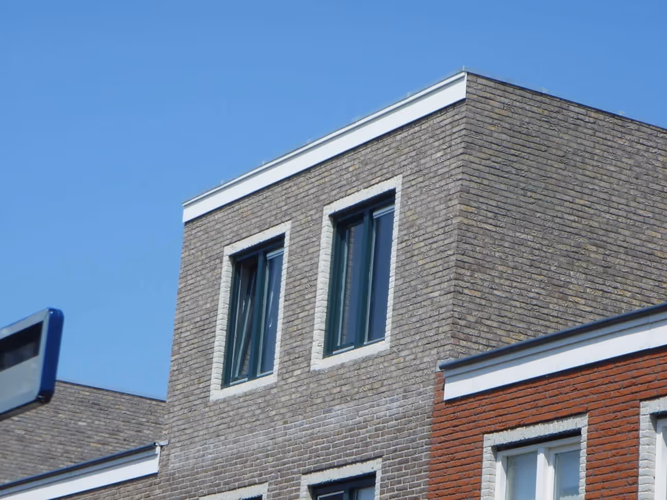 Dakopbouw van een modern gebouw met bruine bakstenen en twee ramen onder een heldere blauwe lucht.