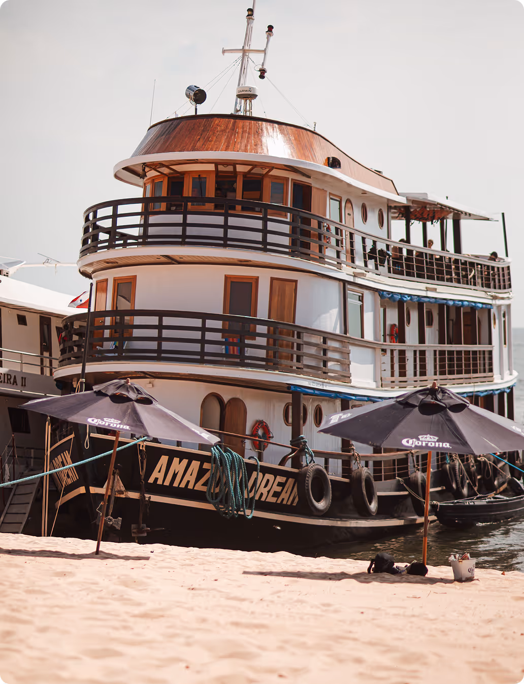 Large white and wooden boat named Amazon Dream docked by a sandy beach with two black Corona-branded umbrellas on the shore.