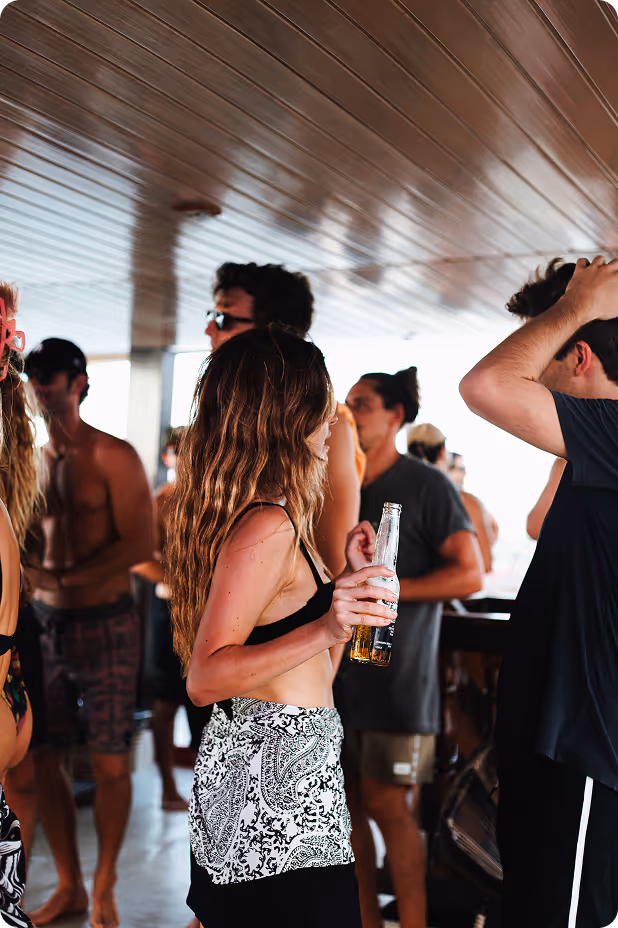 Young woman in a black bikini top and patterned skirt holding a beer bottle at a crowded indoor party.