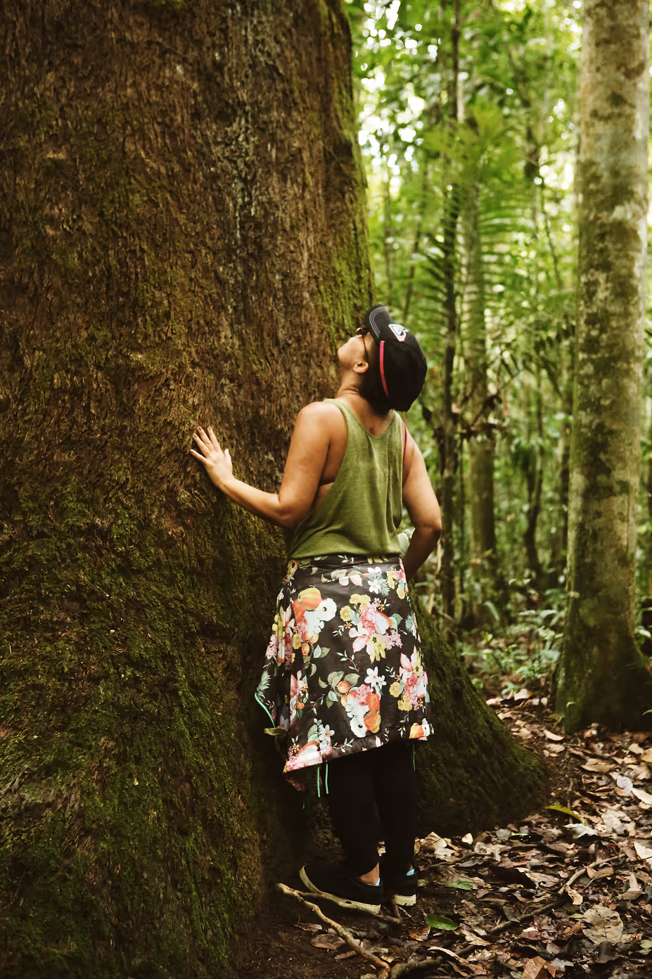Person wearing green tank top and floral skirt touching and looking up at a large moss-covered tree trunk in a dense forest.